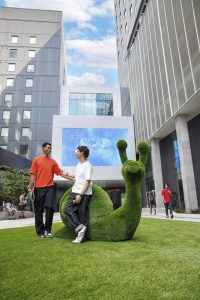 two students standing next to snail-shaped hedge in SCAD Court, giant LED screen in background above