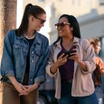 2 femails standing, smiling at each other wearing sunglasses. Female on left in jean jacket. Female on right in layered shirts holding mobile phone.