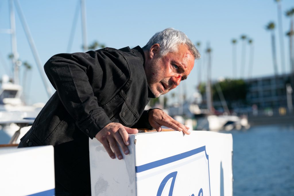 Bosch: Legacy character Harry Bosch leans against the edge of a boat with blood on his face and wearing a black jacket