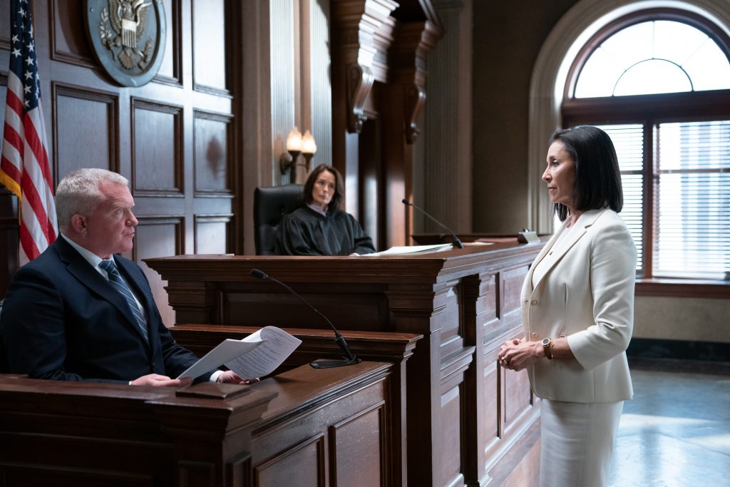 Female in white suit addressing man on the stand in a courtroom. Judge in black robe in the background.
