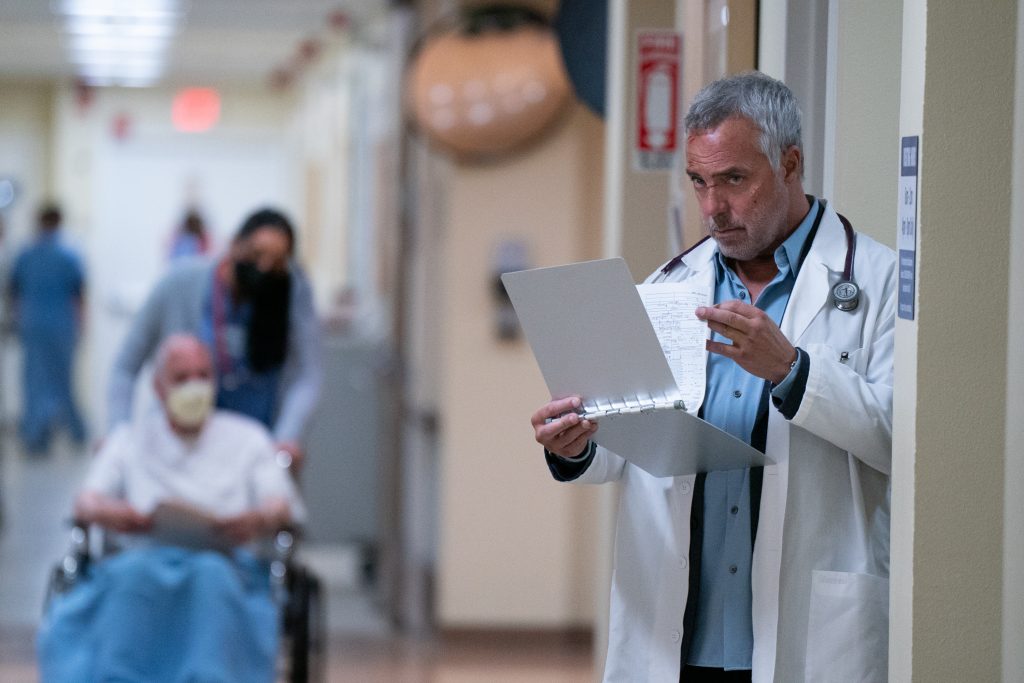Harry Bosch character dressed in doctor's white coat looking at clipboard